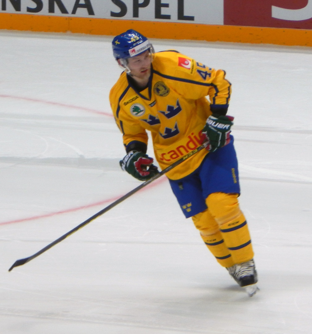 Ein Eishockeyspieler in Helm, Handschuhen und Schlittschuhen hält einen Eishockeyschläger auf dem Eis, mit einer Tafel im Hintergrund.