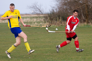 Zwei Personen spielen Hockey auf einem Feld mit Bäumen und Himmel im Hintergrund.