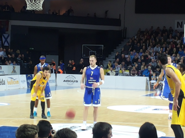 Eine Gruppe von Männern in blauen und gelben Uniformen spielt Basketball auf einem Court mit einem Netz, umgeben von Zuschauern in den Tribünen, während eines 7days Eurocup-Spiels zwischen Fenerbahçe und Real Madrid.