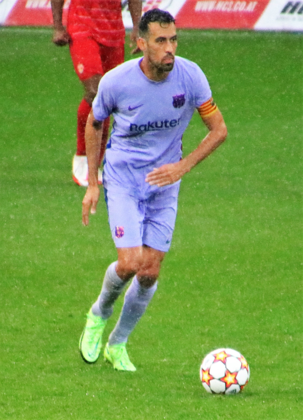 Fußballspieler in Sportkleidung und Fußballschuhen auf einem Feld mit einer Texttafel im Hintergrund.