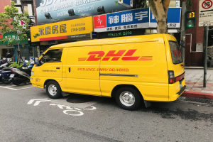 Gelber DHL-Lieferwagen auf der Straße geparkt, umgeben von Gebäuden, Bäumen, Texttafeln, Pfosten, Verkehrsampeln und einer Person auf dem Gehweg.