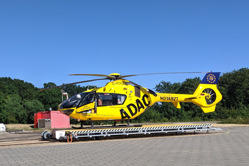 Ein gelber Hubschrauber steht auf einem Flughafen-Asphalt, umgeben von Bäumen und einem klaren blauen Himmel, mit verschiedenen Gegenständen, die mit dem Hubschrauber in Zusammenhang stehen, verstreut.