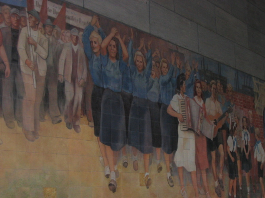 Eine Gruppe von Menschen mit blau gekleideten Mädchen in der Mitte, Musikern auf der rechten Seite, spielenden Kindern in der rechten Ecke und Individuals mit roten Fahnen auf der linken Seite.