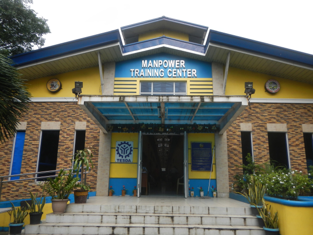 Außenansicht des Manpower Training Centers in Manila, Philippinen, mit Treppen, die zum Eingang führen, flankiert von Topfpflanzen, einem Baum auf der linken Seite und einem Gebäude mit Fenstern, Türen und Schildern unter einem sichtbaren Himmel.
