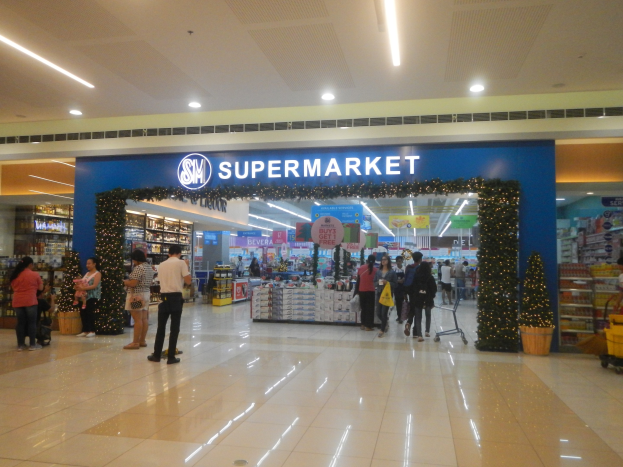 Gruppe von Menschen vor einem Supermarkt mit Einkaufstägern, Taschen, Einkaufswagen, Topfpflanze, Regalen, Schildern und Deckenleuchten.