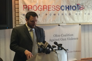 Eine Person steht auf einer Bühne vor einem Tisch mit Mikrofonen, mit einem Banner an der Wand dahinter.