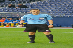 Ein Mann in einem blauen Hemd und schwarzen Shorts steht auf einem Fussballfeld, umgeben von sitzenden und stehenden Menschen, die Fussballschuhe tragen.