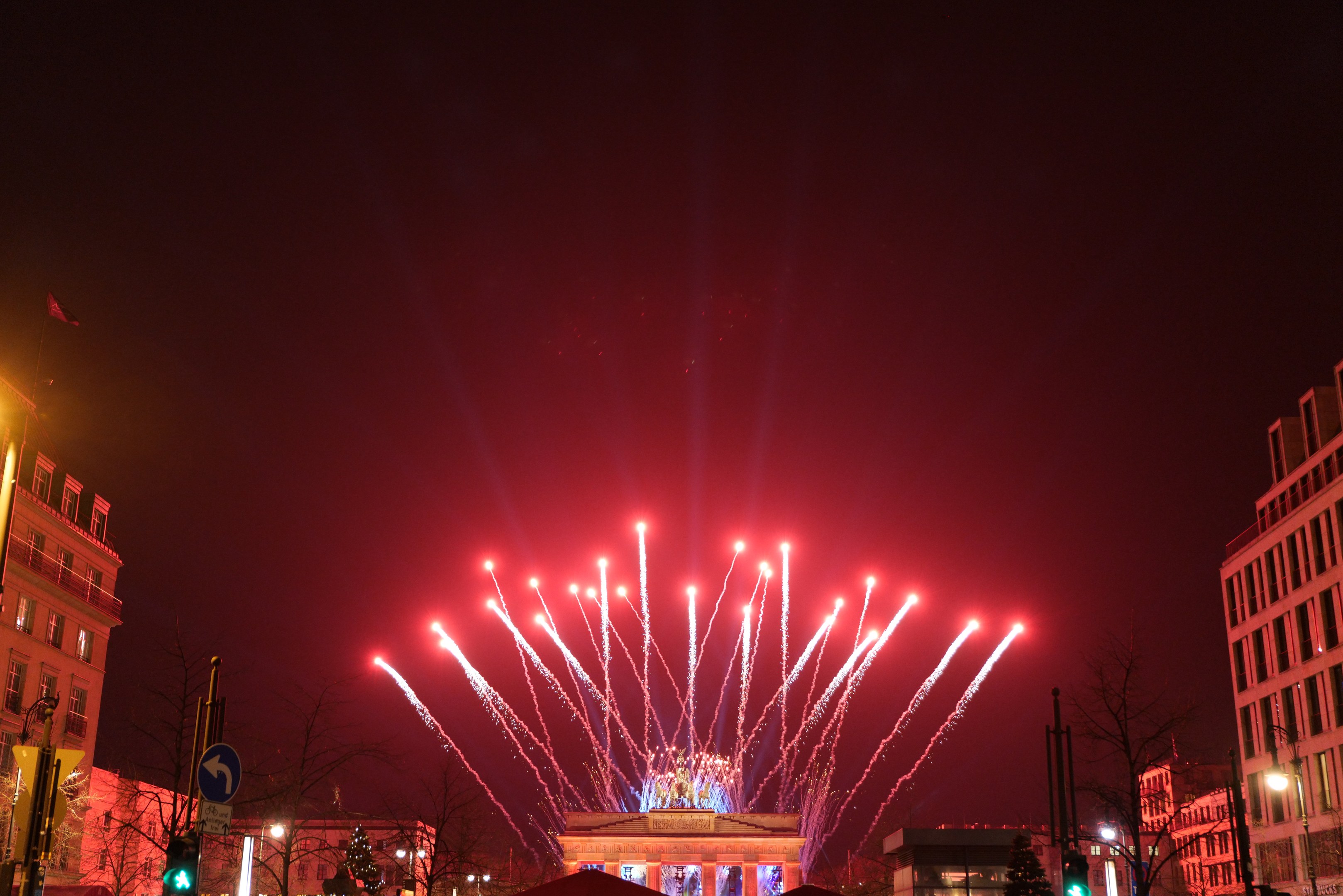 Nachtansicht des Berliner Brandenburger Tors mit Feuerwerk über der beleuchteten Stadtstraße voller Gebäude, Bäume, Pfosten, Verkehrszeichen, Schilder und Fahnen.