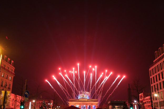 Nachtansicht des Berliner Brandenburger Tors mit Feuerwerk über der beleuchteten Stadtstraße voller Gebäude, Bäume, Pfosten, Verkehrszeichen, Schilder und Fahnen.