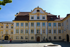 Ein weißes und gelbes Haus mit vielen Glasfenstern, mit zwei älteren Frauen, die auf einem Kopfsteinpflasterweg vor ihm gehen.