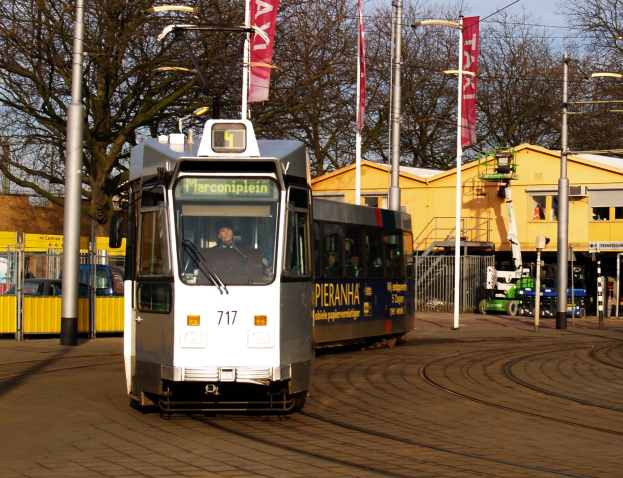 Eine weiße und blaue Straßenbahn auf einer Stadtstraße mit einer Person darin, umgeben von Laternen, Bannern, einem Zaun, Fahrzeugen, Gebäuden, Bäumen und einem klaren blauen Himmel.