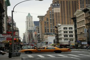 Gelbes Taxi fährt auf einer belebten Stadtstraße mit hohen Gebäuden, Fußgängern auf dem Gehweg, Verkehrsampeln und Texttafeln, unter einem sichtbaren Himmel.