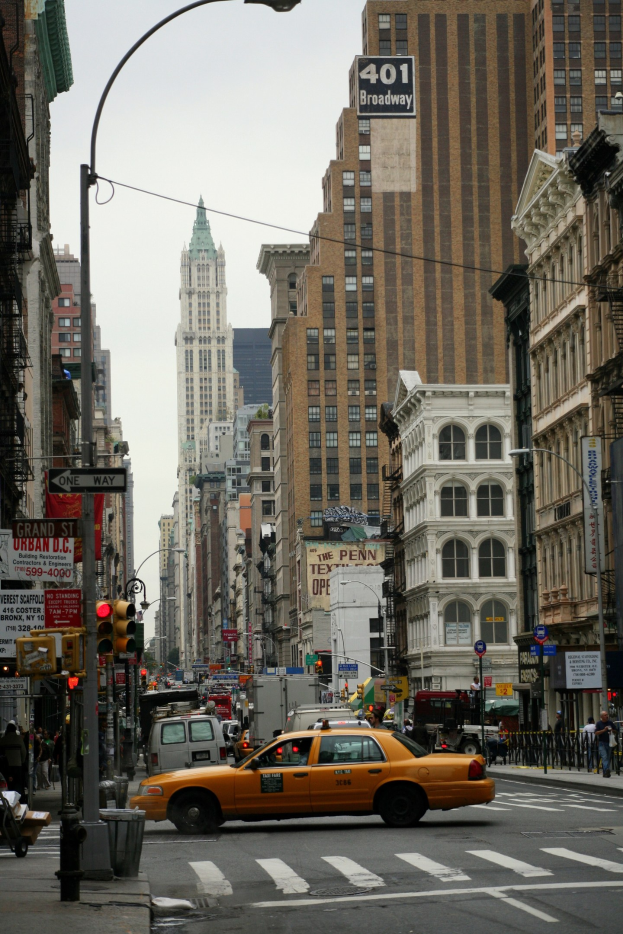 Gelbes Taxi fährt auf einer belebten Stadtstraße mit hohen Gebäuden, Fußgängern auf dem Gehweg, Verkehrsampeln und Texttafeln, unter einem sichtbaren Himmel.
