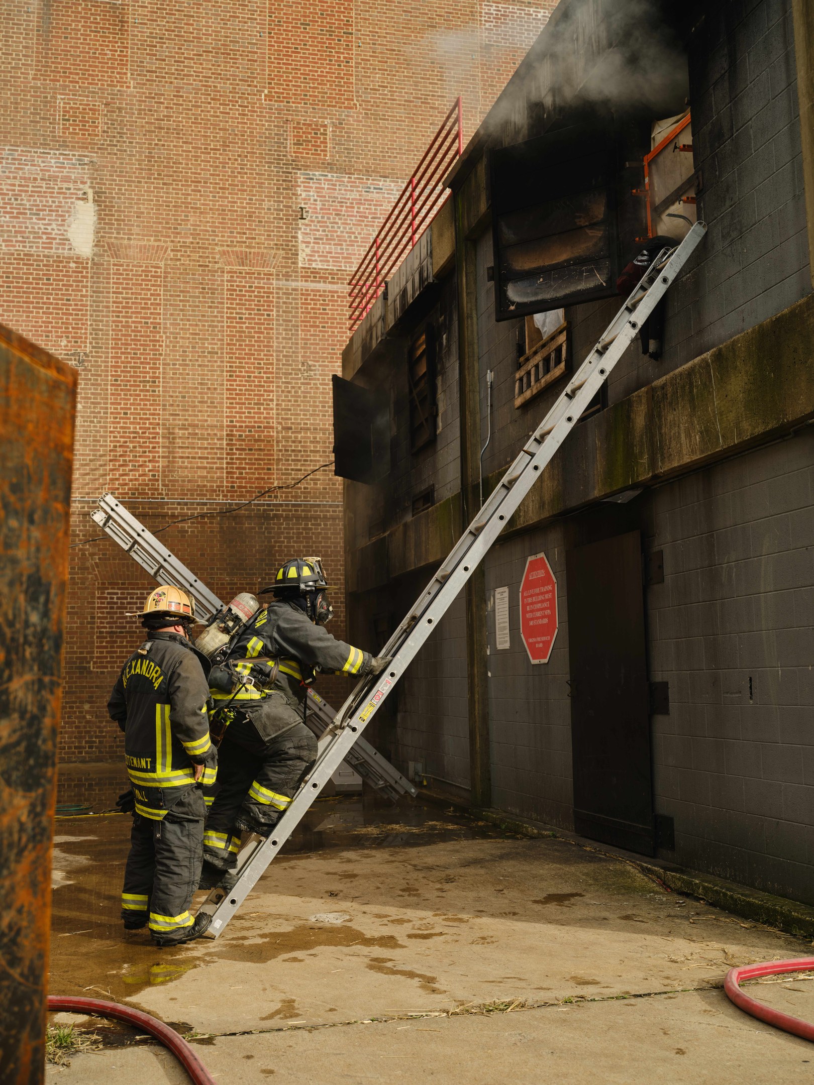 Feuerwehrleute mit Helmen, die eine Leiter hinaufsteigen und auf ein raucherfülltes Gebäude zusteuern, mit einem metallischen Objekt links und Rohren am Boden.