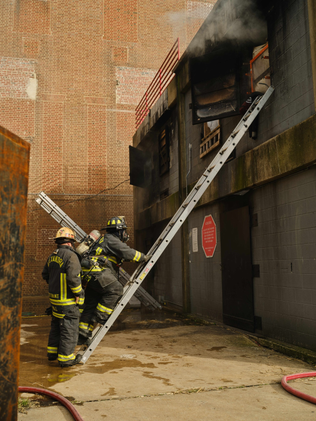 Feuerwehrleute mit Helmen, die eine Leiter hinaufsteigen und auf ein raucherfülltes Gebäude zusteuern, mit einem metallischen Objekt links und Rohren am Boden.