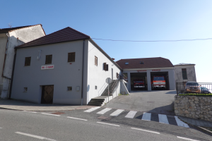 Eine Feuerwache mit einem Feuerwehrauto davor, umgeben von Gebäuden, Fahrzeugen, einem Maschendrahtzaun, Pflanzen, Bäumen und einem Schild, unter einem Himmel.