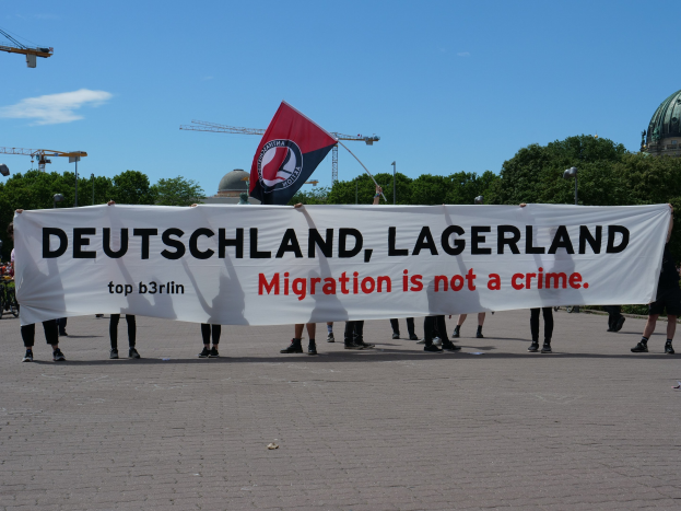Menschen mit einem Banner "Deutschland, Lagerland Migration ist kein Verbrechen" und einer Flagge, mit Straßenlaternen, Bäumen, Kränen, einem Gebäude und einem bewölkten Himmel im Hintergrund.