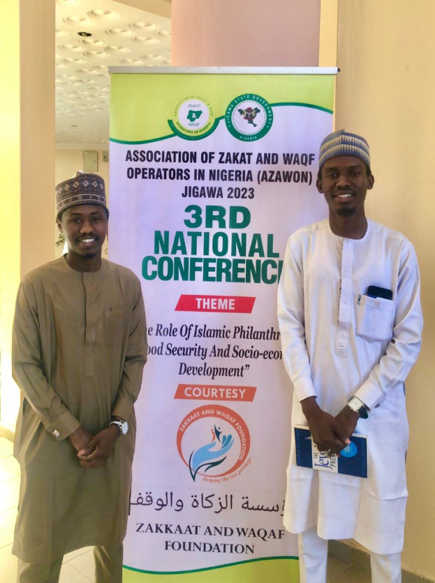Zwei Männer stehen nebeneinander vor einem Banner mit der Aufschrift "3. Nationale Konferenz" mit einer Säule und Deckenbeleuchtung im Hintergrund.