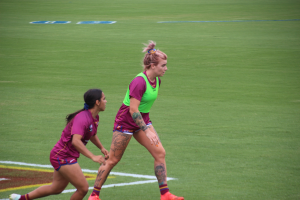 Zwei Frauen in Sportkleidung spielen Fußball auf einem Feld, eine in einem grünen T-Shirt und Shorts, die andere in einem pinken Kleid, beide mit sichtbaren Tattoos an Händen und Füßen.