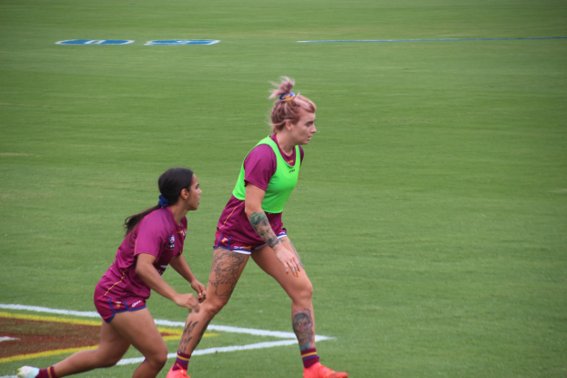 Zwei Frauen in Sportkleidung spielen Fußball auf einem Feld, eine in einem grünen T-Shirt und Shorts, die andere in einem pinken Kleid, beide mit sichtbaren Tattoos an Händen und Füßen.