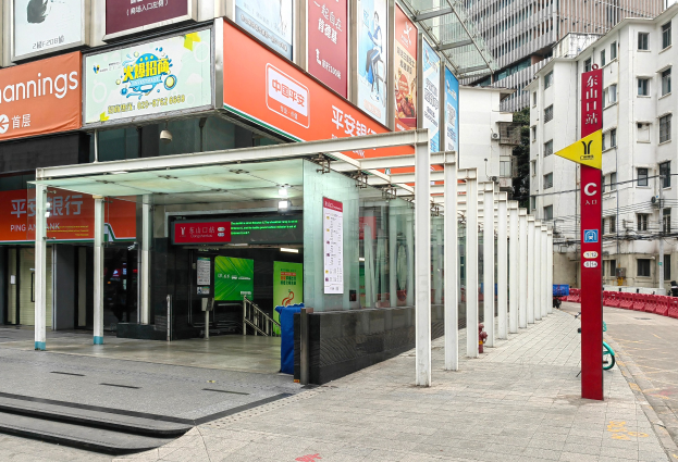 Eine Stadtstraße mit einem Bushaltestellen-Schild, Gebäuden auf beiden Seiten, einer Straße mit einem Mülleimer, roten und gelben Schildern mit Text, Menschen auf dem Gehweg und einer Rolltreppe auf der linken Seite.
