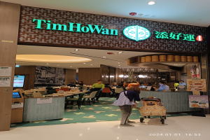 Ein Tim Ho Wan Restaurant in Hong Kong mit Sitzenden und Stehenden, Tischen mit verschiedenen Gegenständen, einer Schautafel, einem Display-Bildschirm, einer Wand mit angeklebten Papieren und einer Decke mit Lampen.