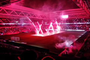 Ein großer Stadion mit Zuschauern, die ein Füßballspiel beobachten, beleuchtet von hellen Lichtern mit Trägern und anderen Gegenständen drumherum und einer Gruppe von Menschen unten, die das Spiel beobachten.