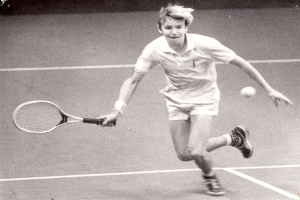 Ein junger Mann in weißer Tennisbekleidung schlägt mit einem Schläger einen Ball auf einem Tennisplatz, mit entschlossener Miene.