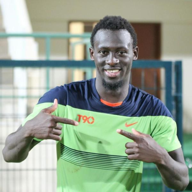 Ein Mann in einem grün-blauen Hemd, der lachend in die Kamera zeigt und im Hintergrund eine undeutliche Brüstung, Wand und Tür zu sehen sind.