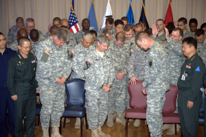 Eine Gruppe von Menschen in militärischer Kleidung steht zwischen roten und blauen Stühlen, mit Flaggen im Hintergrund.