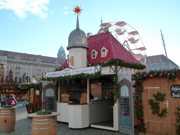 Ein kleines rotes Gebäude mit einem Riesenrad davor, umgeben von festlichen Oktoberfest-Dekorationen und Menschen, vor einer Kulisse aus Gebäuden, Bäumen und einem bewölkten Himmel.