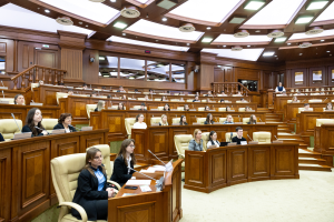 Eine große Gruppe von Menschen sitzt an Tischen in einem Raum, ausgestattet mit Mikrofonen und Papieren, an einer Sitzung des griechischen Parlaments teilnehmend, mit Treppen und Geländern im Hintergrund und Deckenlampen.