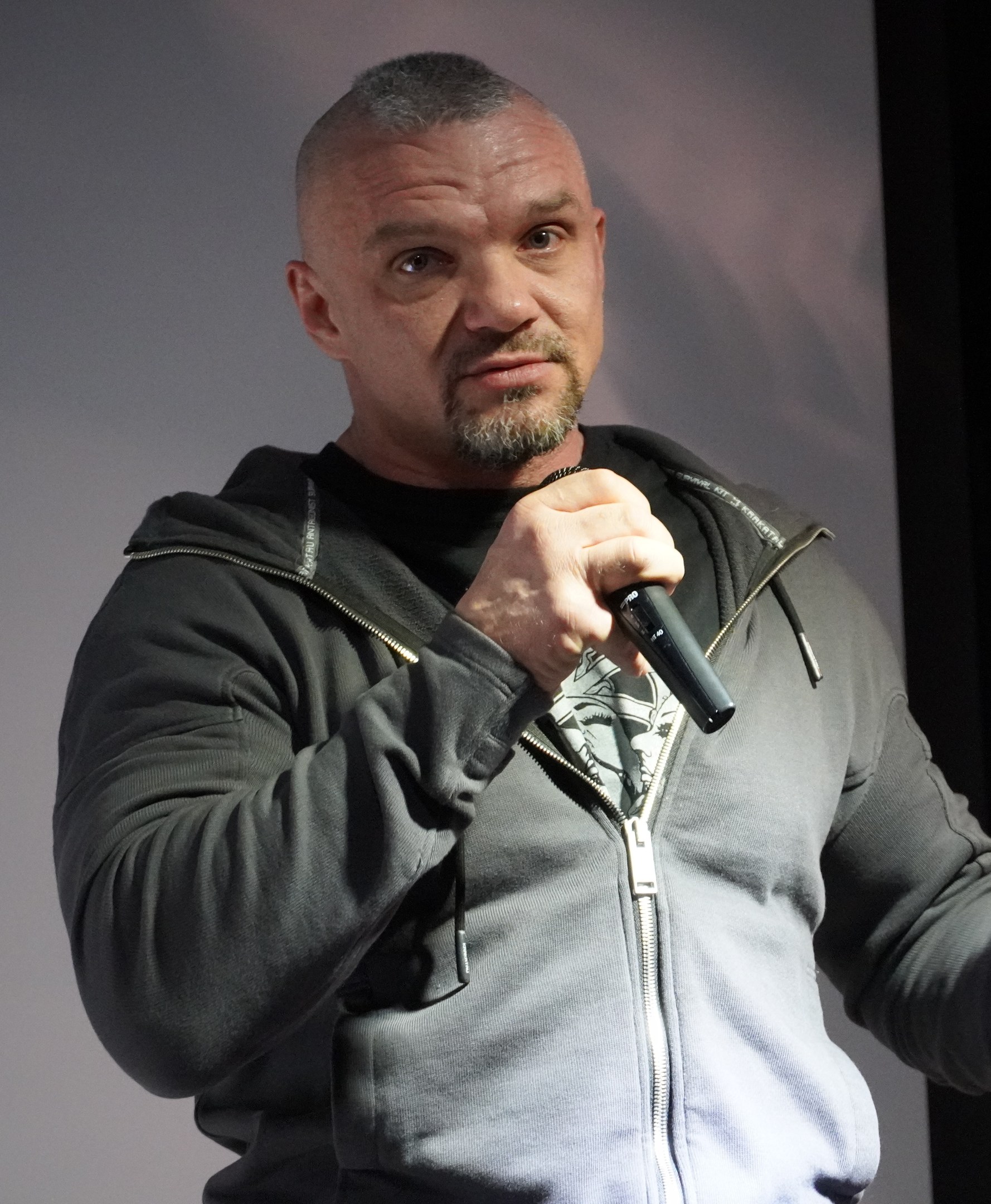 Ein Mann in einer schwarzen Kapuzenjacke und einem schwarzen Anzug mit einem weißen Hemd, glattem Haar und einem selbstbewussten Ausdruck, der ein Mikrofon vor einer Wand hält.