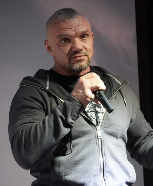 Ein Mann in einer schwarzen Kapuzenjacke und einem schwarzen Anzug mit einem weißen Hemd, glattem Haar und einem selbstbewussten Ausdruck, der ein Mikrofon vor einer Wand hält.