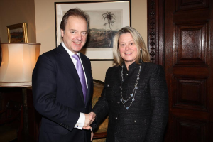 Eine Frau und ein Mann in einem Anzug lächeln, schütteln sich die Hände und sehen sich in einem Raum mit einer Wand, Fotorahmen, einer Lampe, einer Tür und ein paar Gegenständen an.