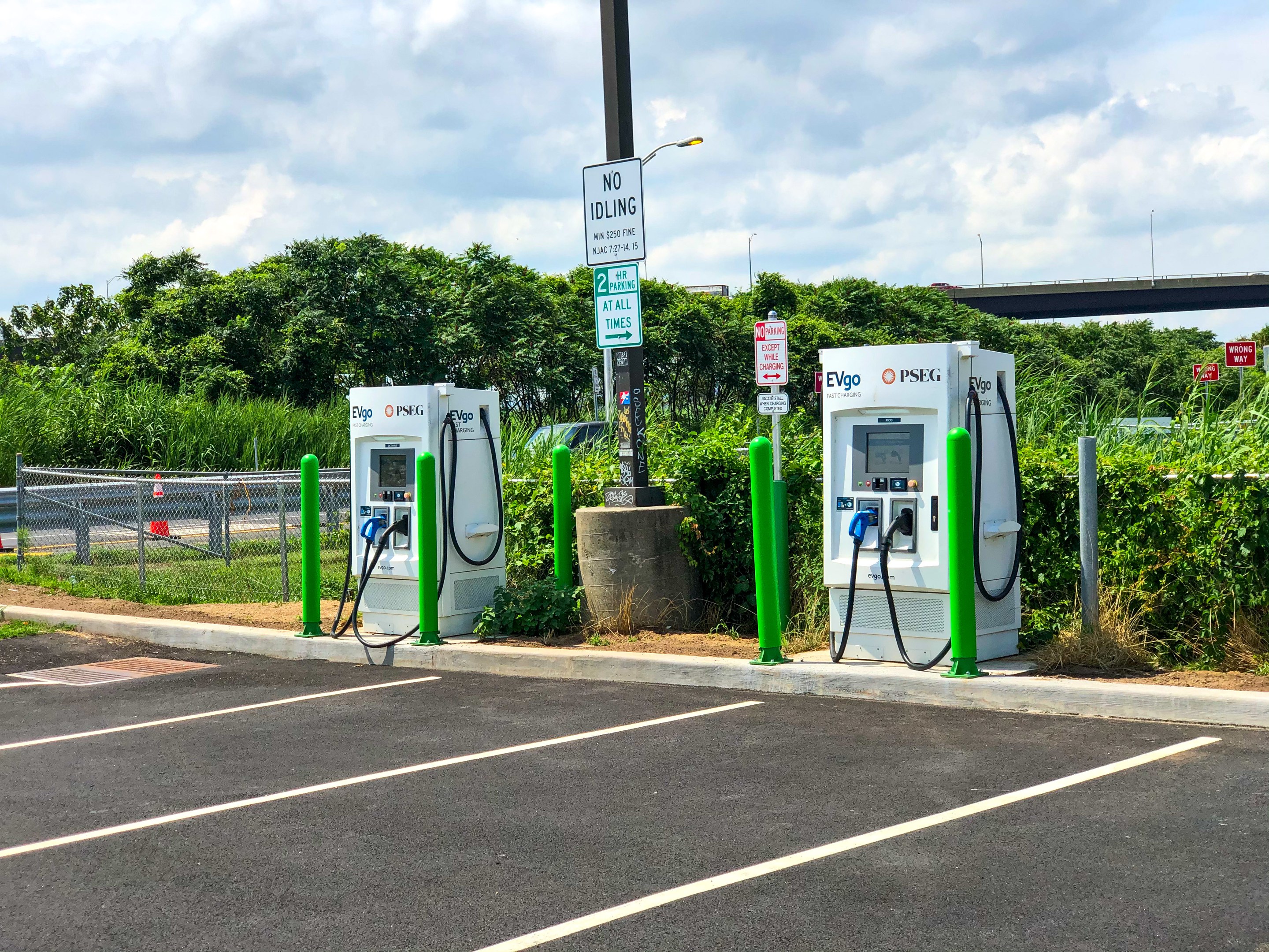 Elektrisches Fahrzeug-Ladestation auf einem Parkplatz mit umgebenen Infrastruktur, Grünflächen und einer Brücke im Hintergrund bei bewölktem Himmel.