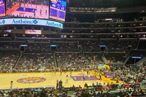 Ein Basketballspiel wird in einer großen, hell erleuchteten Arena mit Zuschauern in den Rängen, Treppen, Geländern, einem großen Bildschirm und Texttafeln am Rand gespielt.