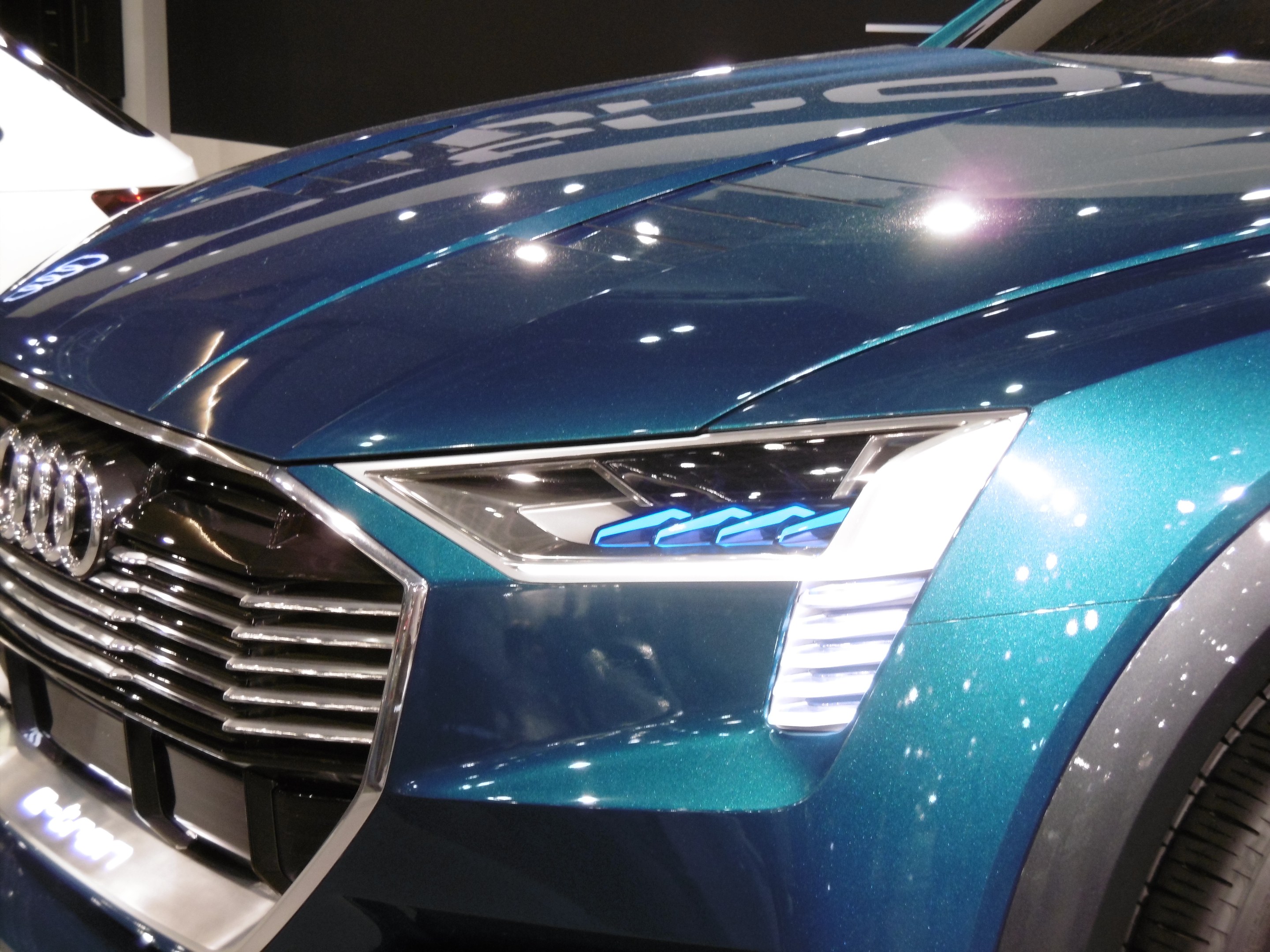 Blauer Audi e-tron Conceptwagen auf einer Automesse zu sehen, mit einer schlanken Gestaltung mit einer gebogenen Motorhaube, einem großen Kühlergrill, winkligen Scheinwerfern und sportlichen Rädern, vor einem weißen Auto und einer schwarzen Wand positioniert.