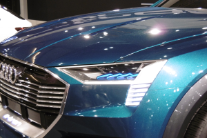 Blauer Audi e-tron Conceptwagen auf einer Automesse zu sehen, mit einer schlanken Gestaltung mit einer gebogenen Motorhaube, einem großen Kühlergrill, winkligen Scheinwerfern und sportlichen Rädern, vor einem weißen Auto und einer schwarzen Wand positioniert.