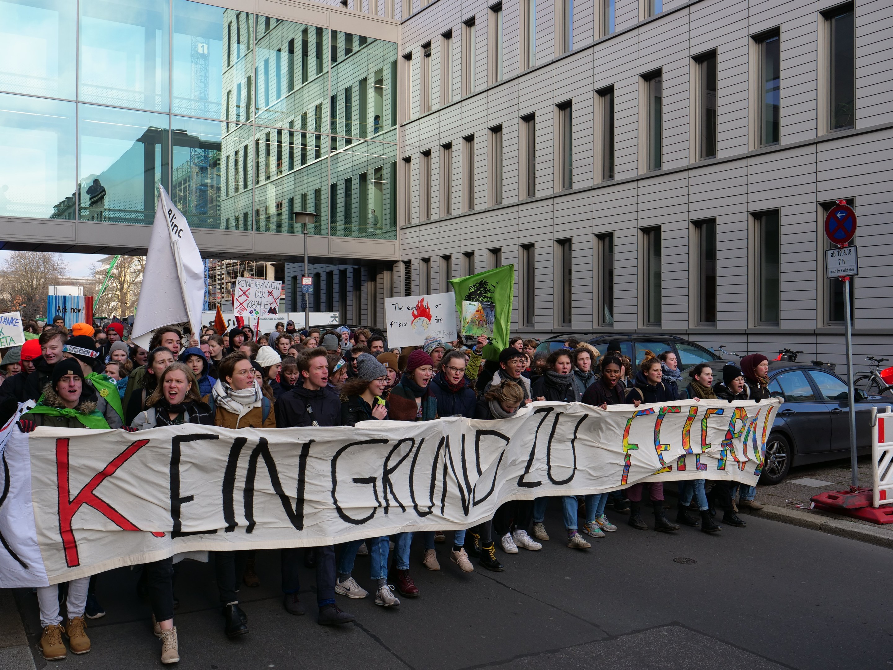 Protestierende mit Transparent "Kein Sparen auf Kosten der Armen" auf einer Straße mit Gebäuden, Bäumen und Fahrzeugen im Hintergrund bei klarem blauem Himmel.