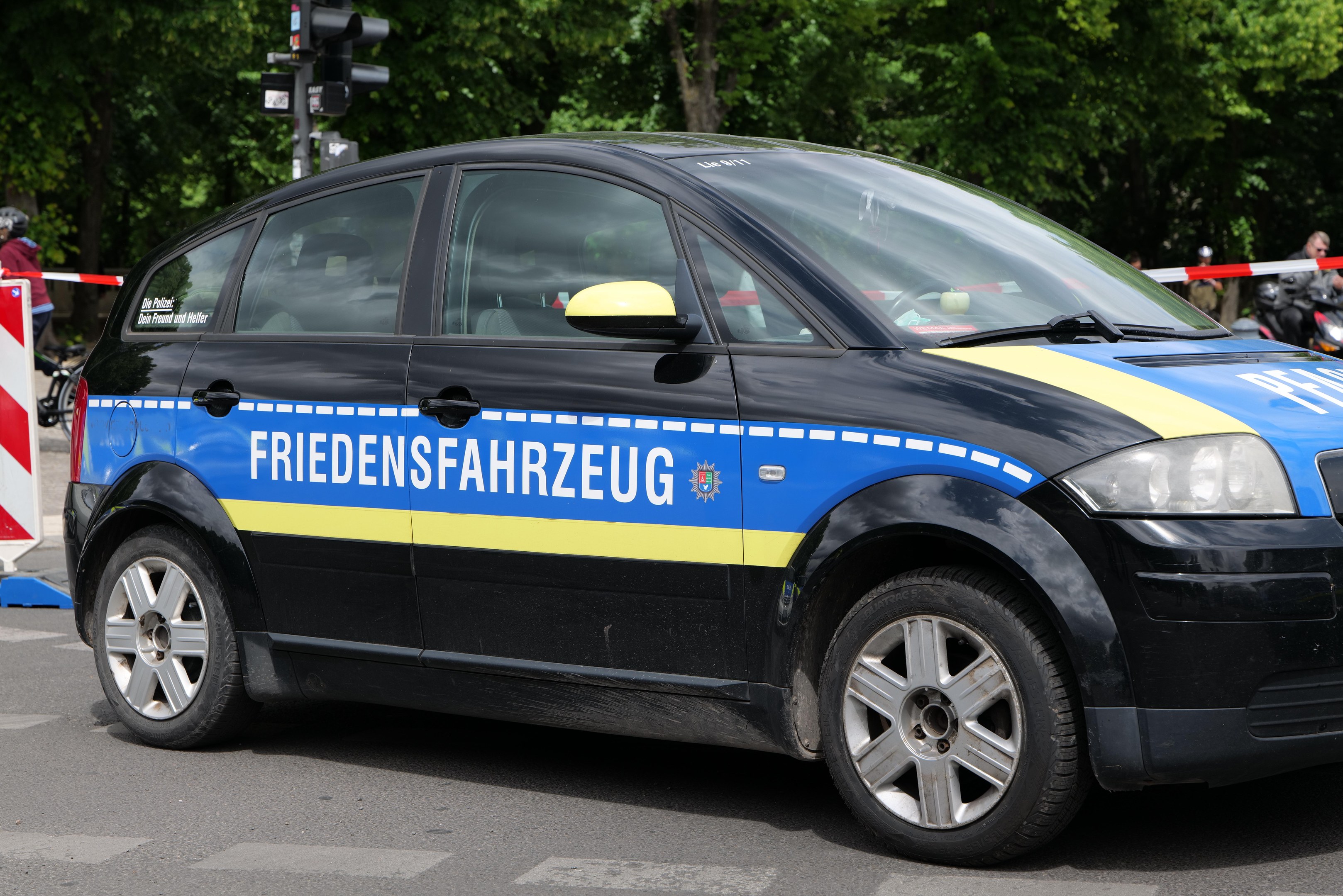 Polizeifahrzeug am Straßenrand mit Text an der Seite, mehrere Personen in der Nähe und Verkehrszeichen mit Bäumen im Hintergrund.