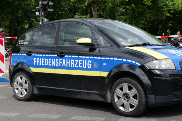 Polizeifahrzeug am Straßenrand mit Text an der Seite, mehrere Personen in der Nähe und Verkehrszeichen mit Bäumen im Hintergrund.