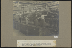 Drei Frauen arbeiten in einer Textilfabrik, stehen zwischen Maschinen, Pfosten und unter Deckenlampen, mit Text am unteren Bildrand.