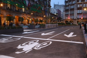 Eine Stadtstraße mit einem Fahrradstreifen, Gebäuden, Straßenlaternen, Verkehrsschildern, Menschen auf dem Gehweg, Bänken, Pflanzen, Bäumen, dekorativen Elementen und einem klaren blauen Himmel.