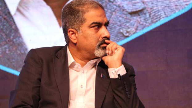 Ein Mann in Anzug und Krawatte sitzt auf einer Bühne vor einem Bildschirm und denkt nach.