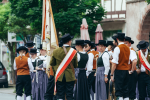 Gruppe von Menschen in traditioneller bayerischer Tracht, die eine Straße entlangmarschieren, einige spielen Instrumente und tragen Fahnen, mit Bäumen, Gebäuden und einem Auto im Hintergrund.