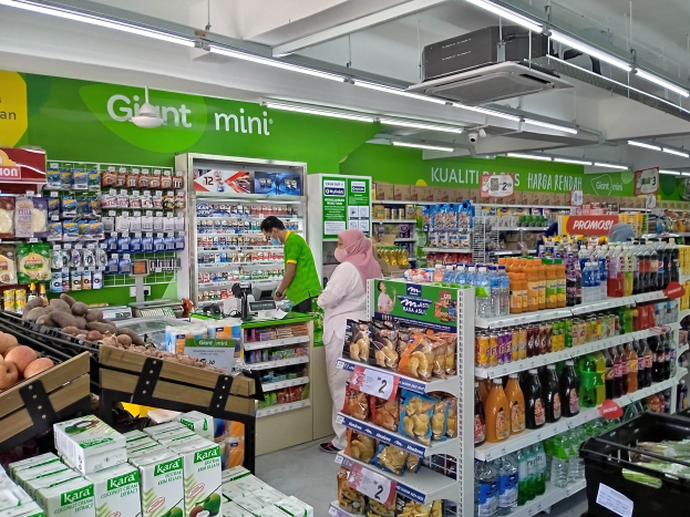 Ein hell erleuchteter Mini-Supermarktgang mit Flaschen, Kartons, Obst und verpackten Lebensmitteln auf Regalen, mit Deckenleuchten, Texttafeln und einem Projektor, der die Eröffnung des Ladens in Malaysia markiert.