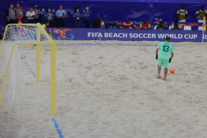 Ein Mann schießt einen Fußball auf einem sandigen Strand mit einem Tor auf der linken Seite und einem Banner mit Text im Hintergrund, umgeben von Zuschauern, einige halten Kameras.
