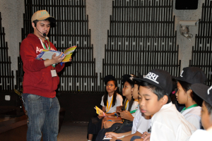 Ein Mann in einer maronenfarbenen Kapuzenjacke und Jeans hält ein Buch und spricht in ein Mikrofon, mit ein paar Kindern in weißen T-Shirts und schwarzen Mützen auf der rechten Seite und einer Kamera an der Wand im Hintergrund.