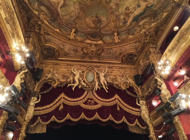 Theaterinnenraum mit roter Gardine, Kronleuchtern, Deckenplastiken, Lichtern und einem Gemälde, das eine Bühne mit einigen Personen am Boden zeigt.